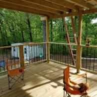 A wooden deck overlooks a forest and a small white building, featuring two orange folding chairs and metal railings.