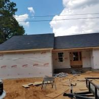 A partially constructed house exterior with dark roof shingles, white house wrap siding, and a wooden front door frame.