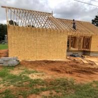 A construction site showing a house frame under development with wood wall panels and an exposed roof truss system.