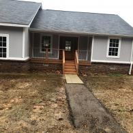 Gray house with a brown stone foundation, a wooden porch with steps, and a dirt yard with a narrow walkway to the entry.