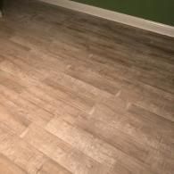 A view of light-colored, wood-grain plank flooring installed against a white baseboard and a dark green wall.