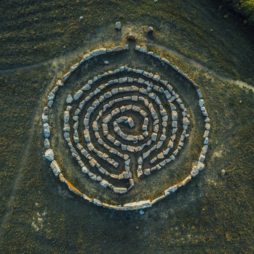 Aerial view of a circular stone labyrinth on a grassy hillside.