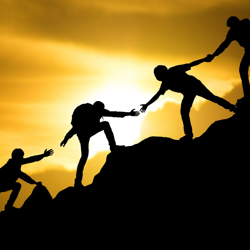 Silhouetted people climbing a mountain, helping each other against a golden sunset.