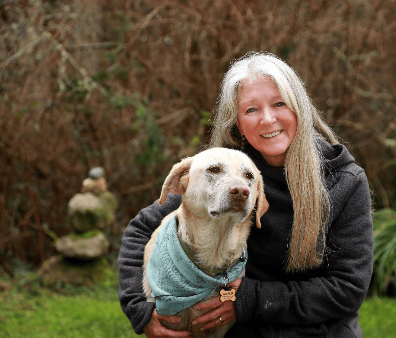 Woman smiling, holding a yellow Labrador wearing a blue scarf, outdoors by a small stacked rock formation.
