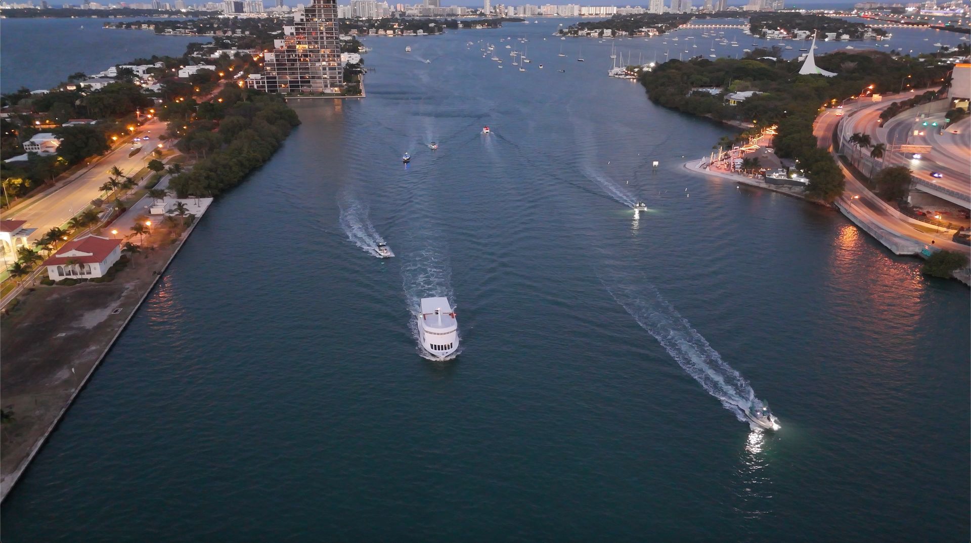 Drone view of a Miami sightseeing sunset cruise boat sailing through Biscayne Bay with evening skyline lights and waterway traffic.