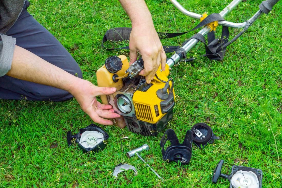 A person works on repairing a yellow string trimmer on a green grassy lawn.