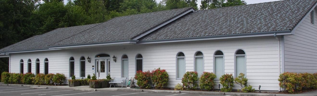 White building with arched windows and a dark shingled roof, viewed from the front amid landscaping.