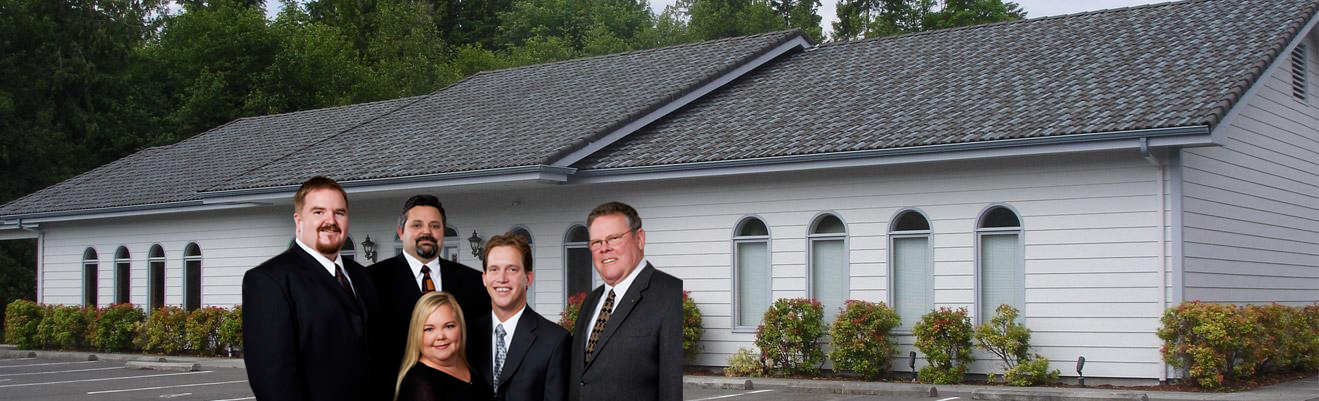 Four people in suits stand outdoors in front of a white building with arched windows and a dark shingled roof