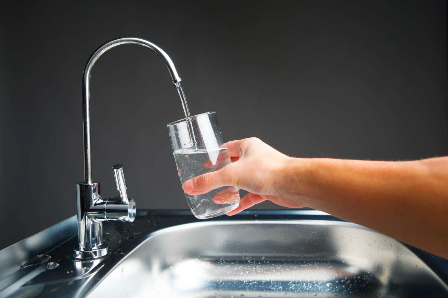 Hand filling a glass with clear water from a kitchen faucet over a stainless-steel sink