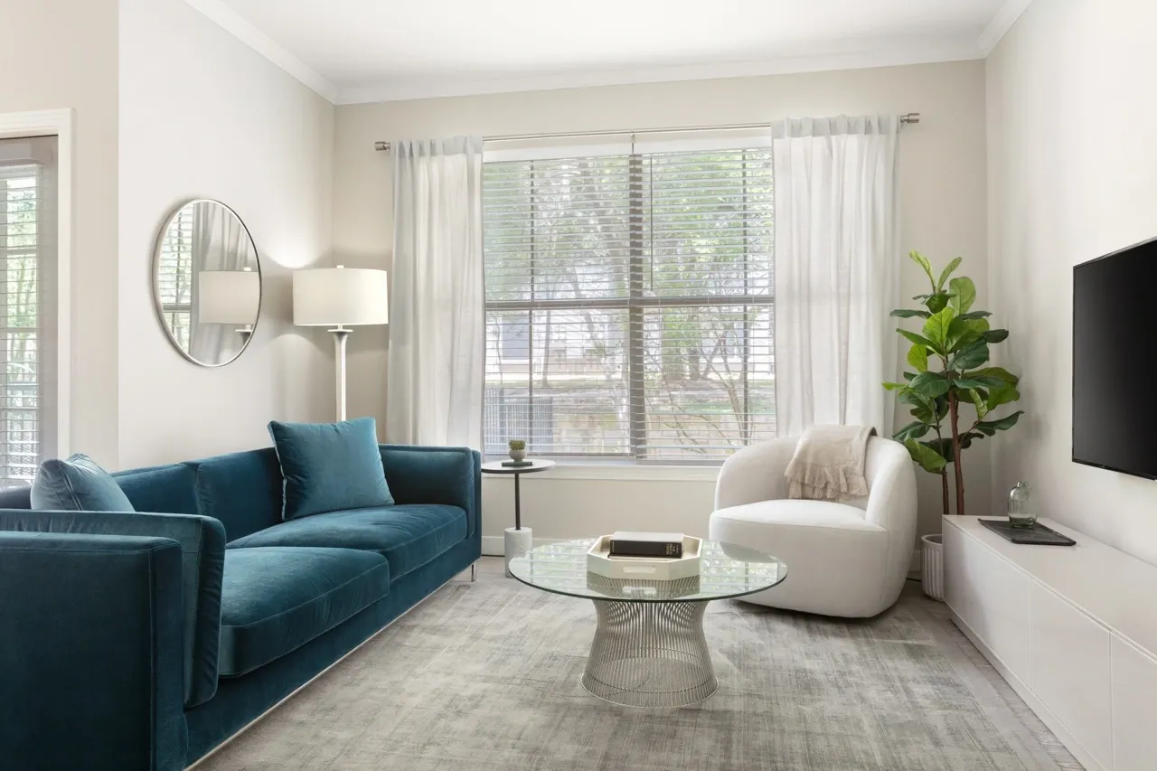 A modern living room with a teal velvet sofa, a white armchair, a glass coffee table, and a window.