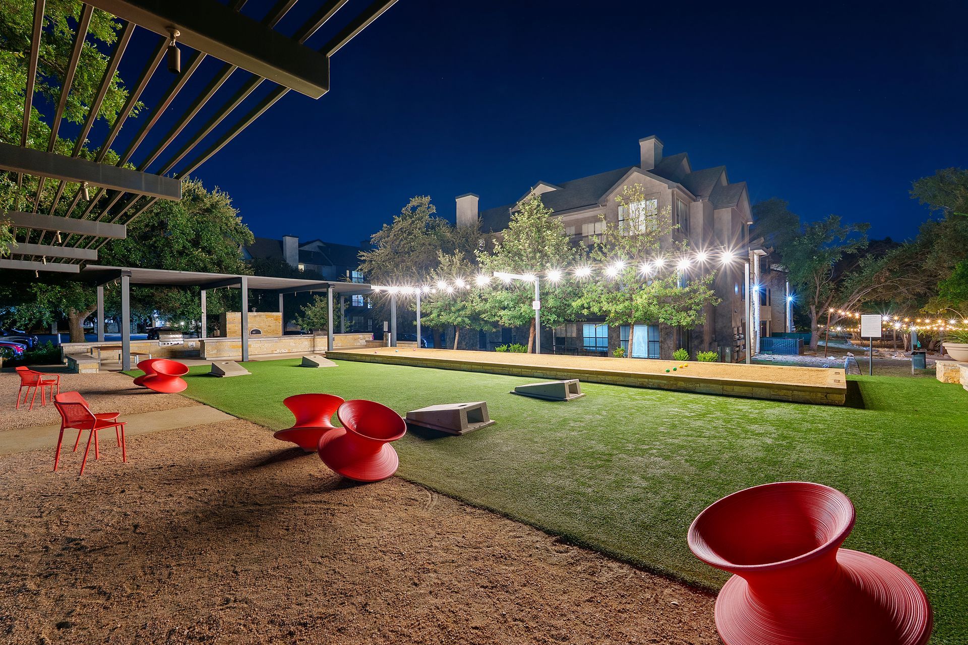 Modern outdoor courtyard at night with red chairs, lit trees, and a grassy lawn