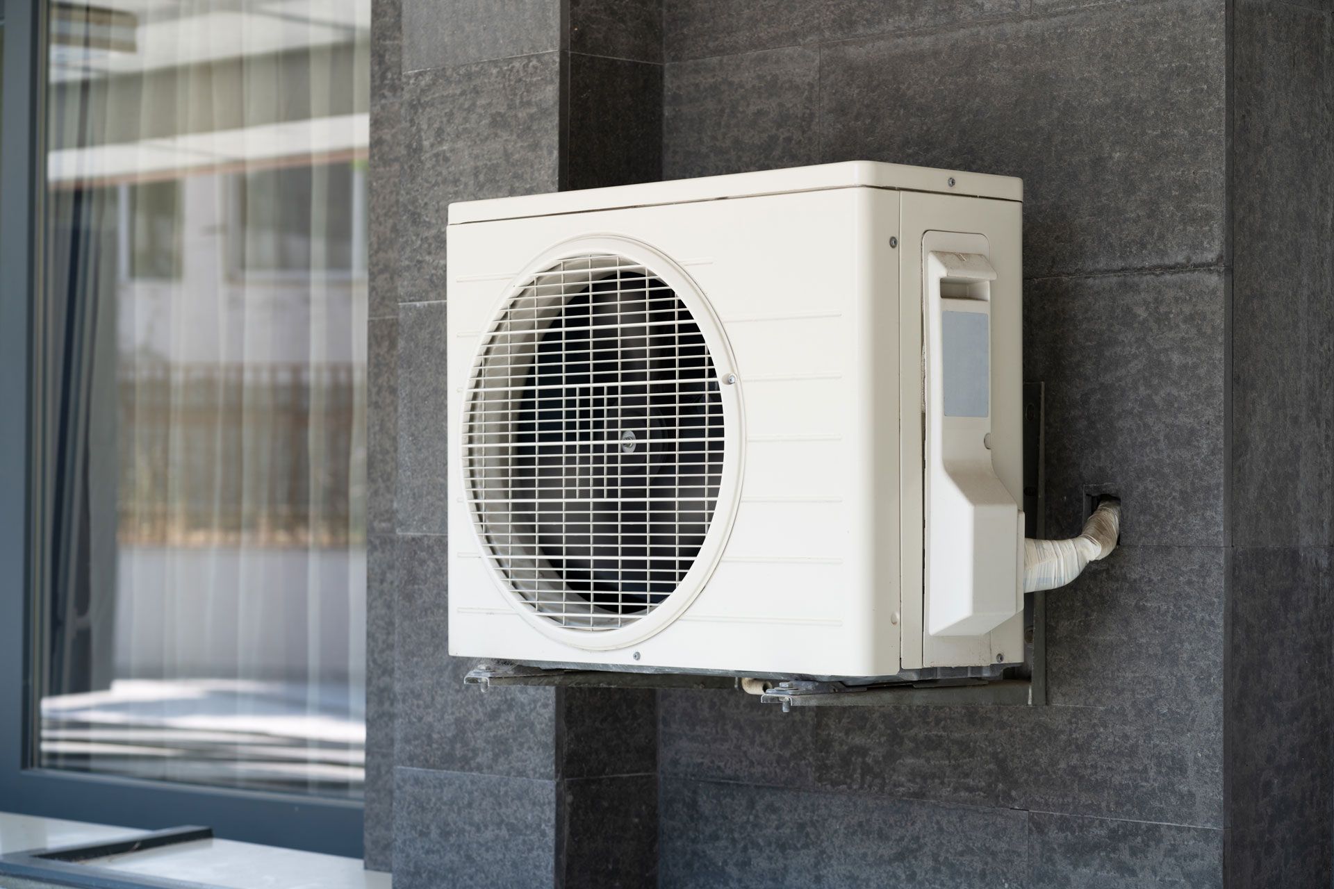 White outdoor air conditioner unit mounted on a dark wall beside a window