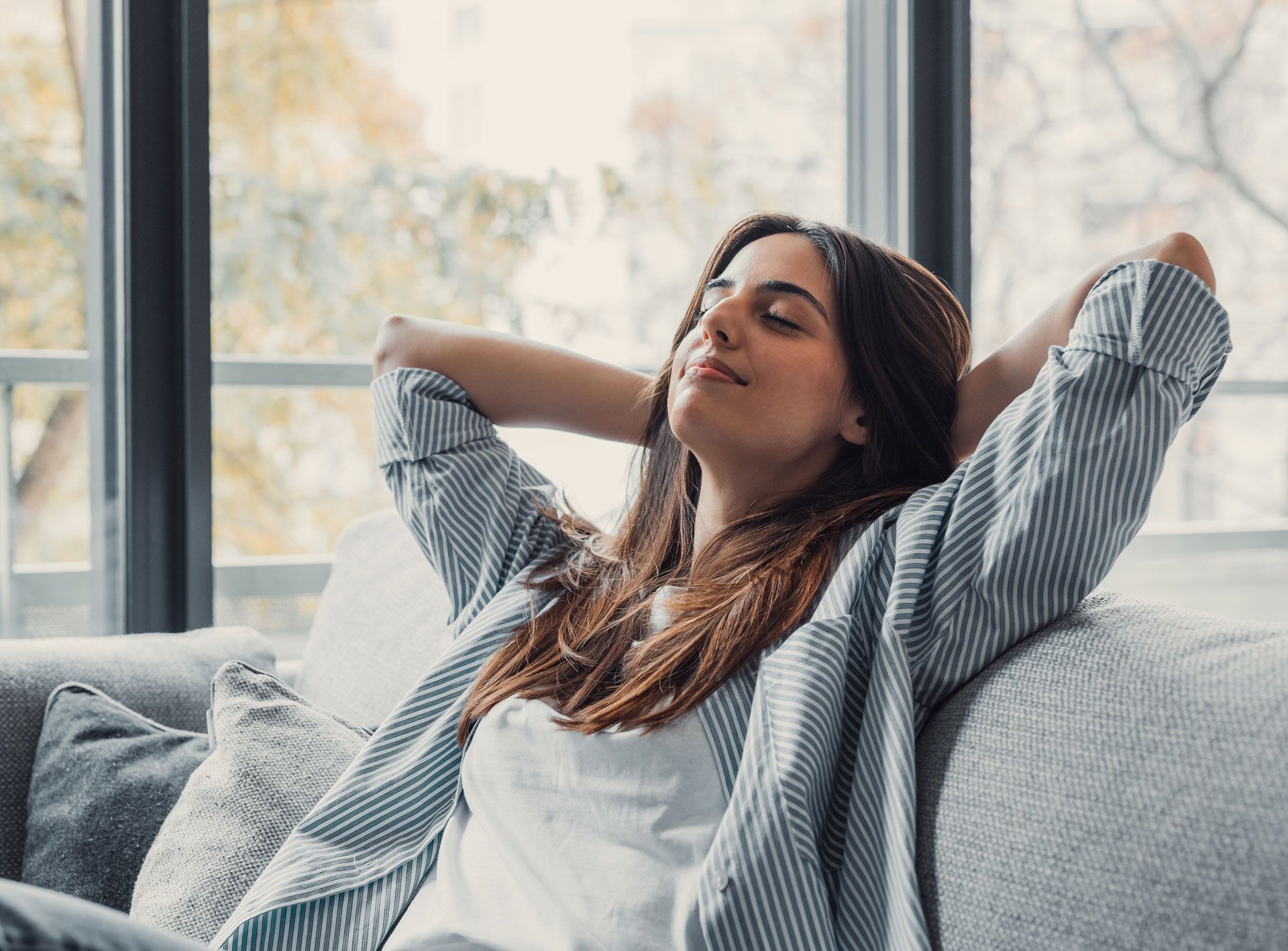 Woman relaxing on a couch with eyes closed, hands behind head, by a bright window