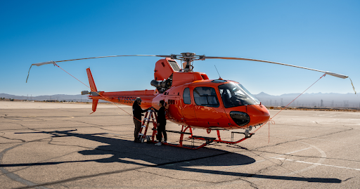 Technicians performing maintenance on an orange Airbus H125 helicopter on a desert airfield in Nevada, illustrating the importance of choosing a certified aircraft repair station.