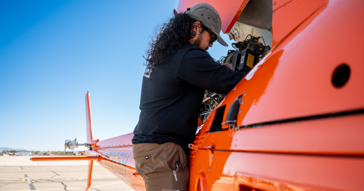 Aviation maintenance technician inspecting equipment inside an open panel of an orange helicopter.
