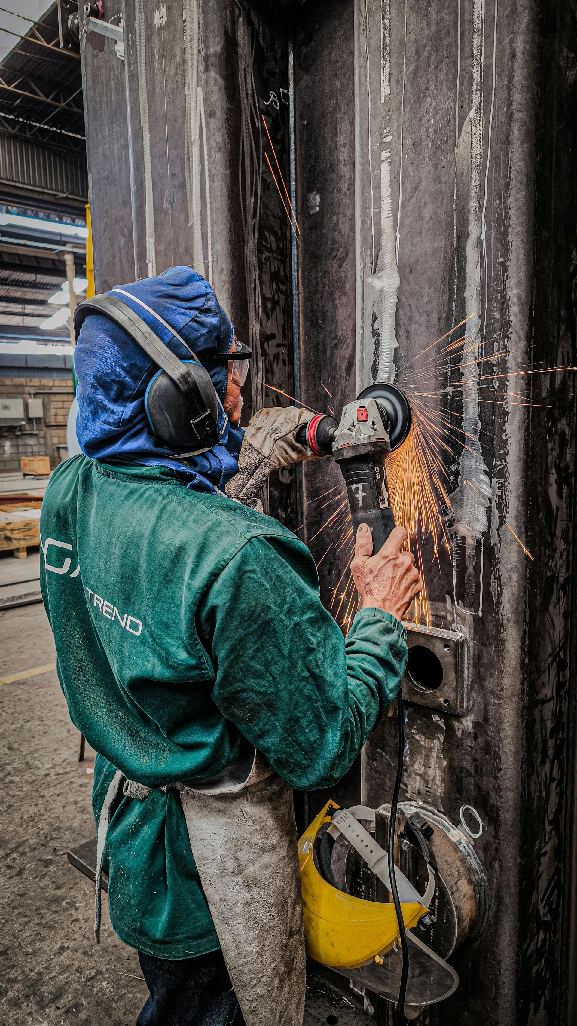 Worker in green coveralls and blue helmet using a power tool on a metal wall, with sparks flying.