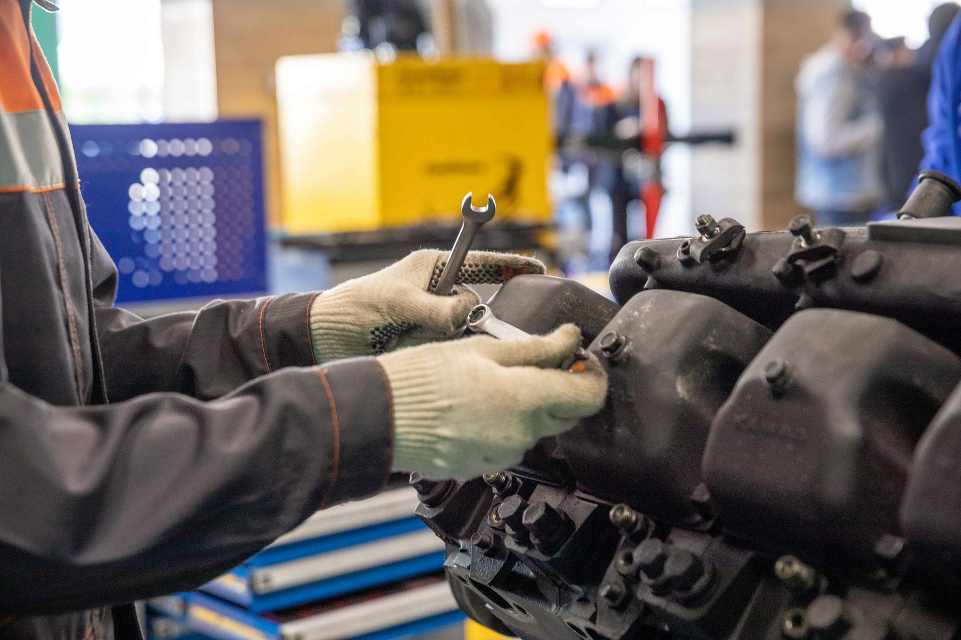 Gloved hands working on a car engine in a workshop, with tools and machinery in the background