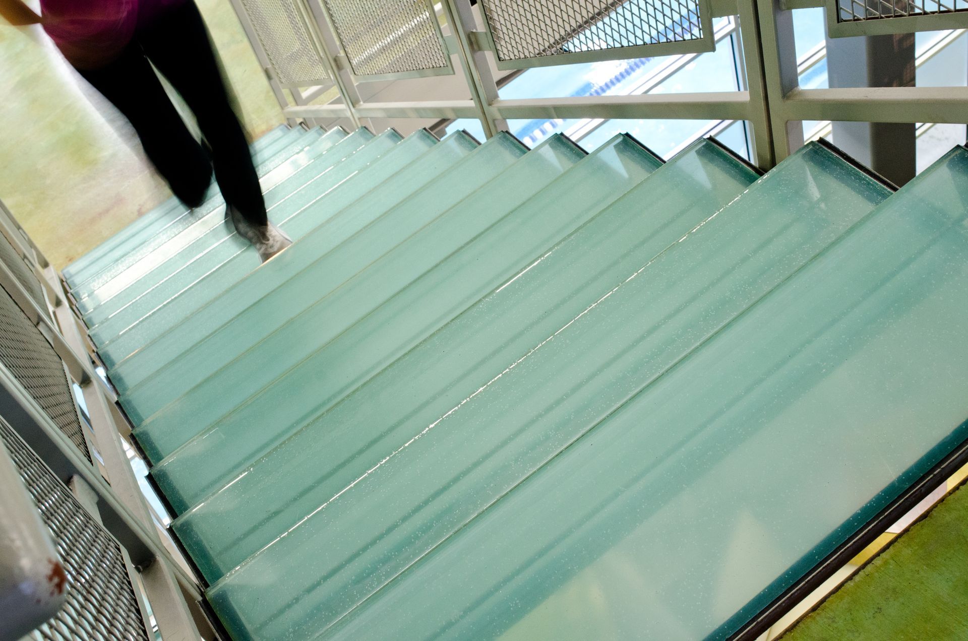 Person walking down glass stairs. Blue-green glass steps, metal railings, modern architecture.