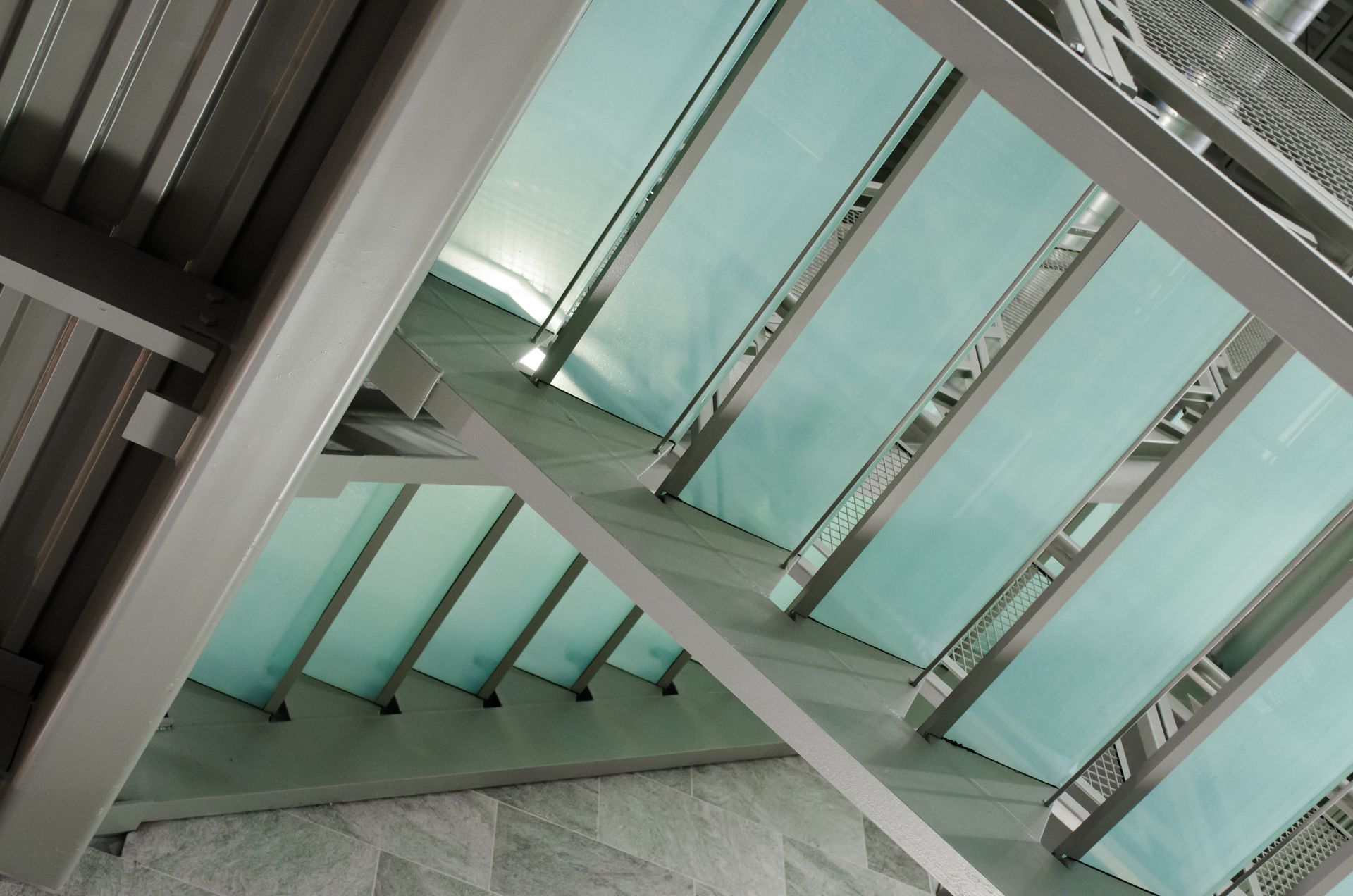 Metal beams support a turquoise-tinted roof, viewed from below. Sunlight streams through.