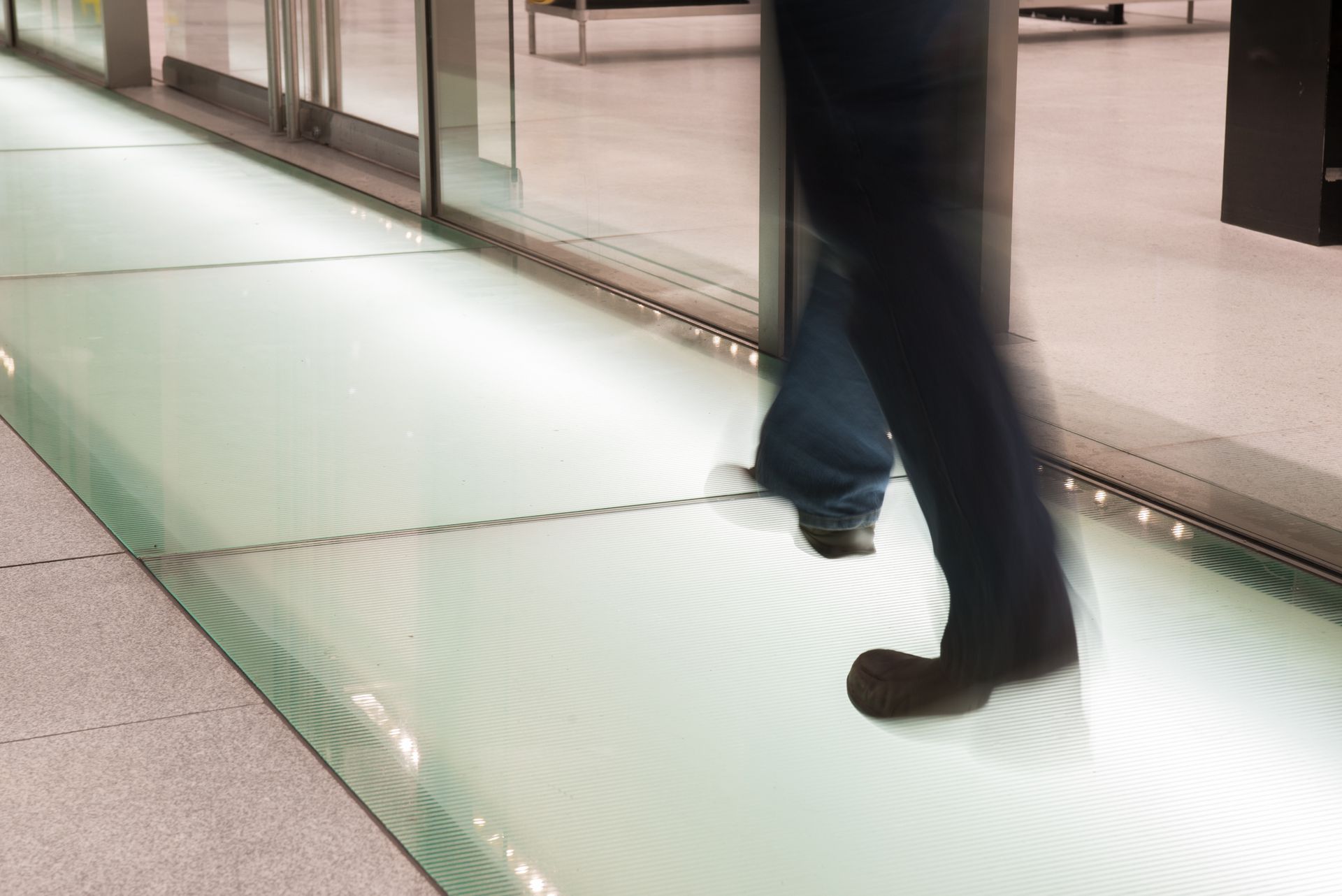 Person walking on a glass floor with light panels, near automatic sliding doors.