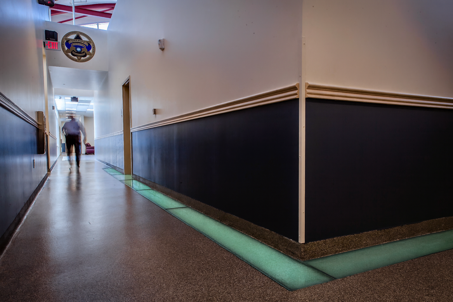 Long hallway with a person walking; blue-walled section, light-filled flooring, and wall decoration with flags.