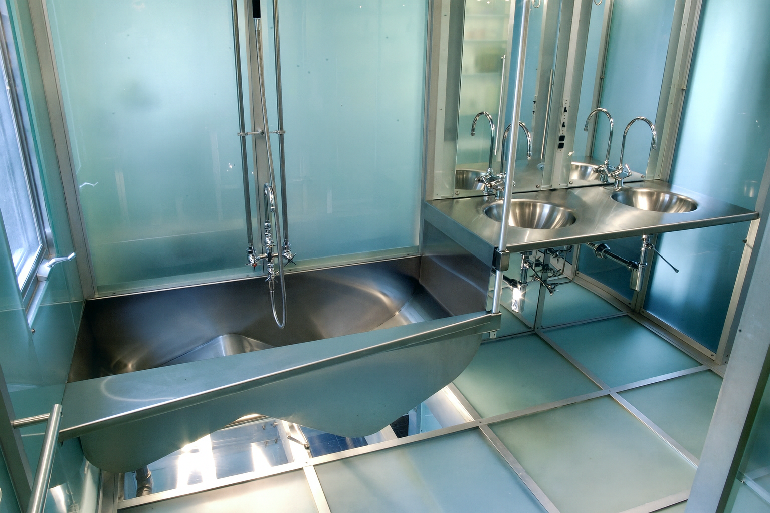Modern bathroom with stainless steel sink, shower, and multiple faucets; walls are frosted glass.