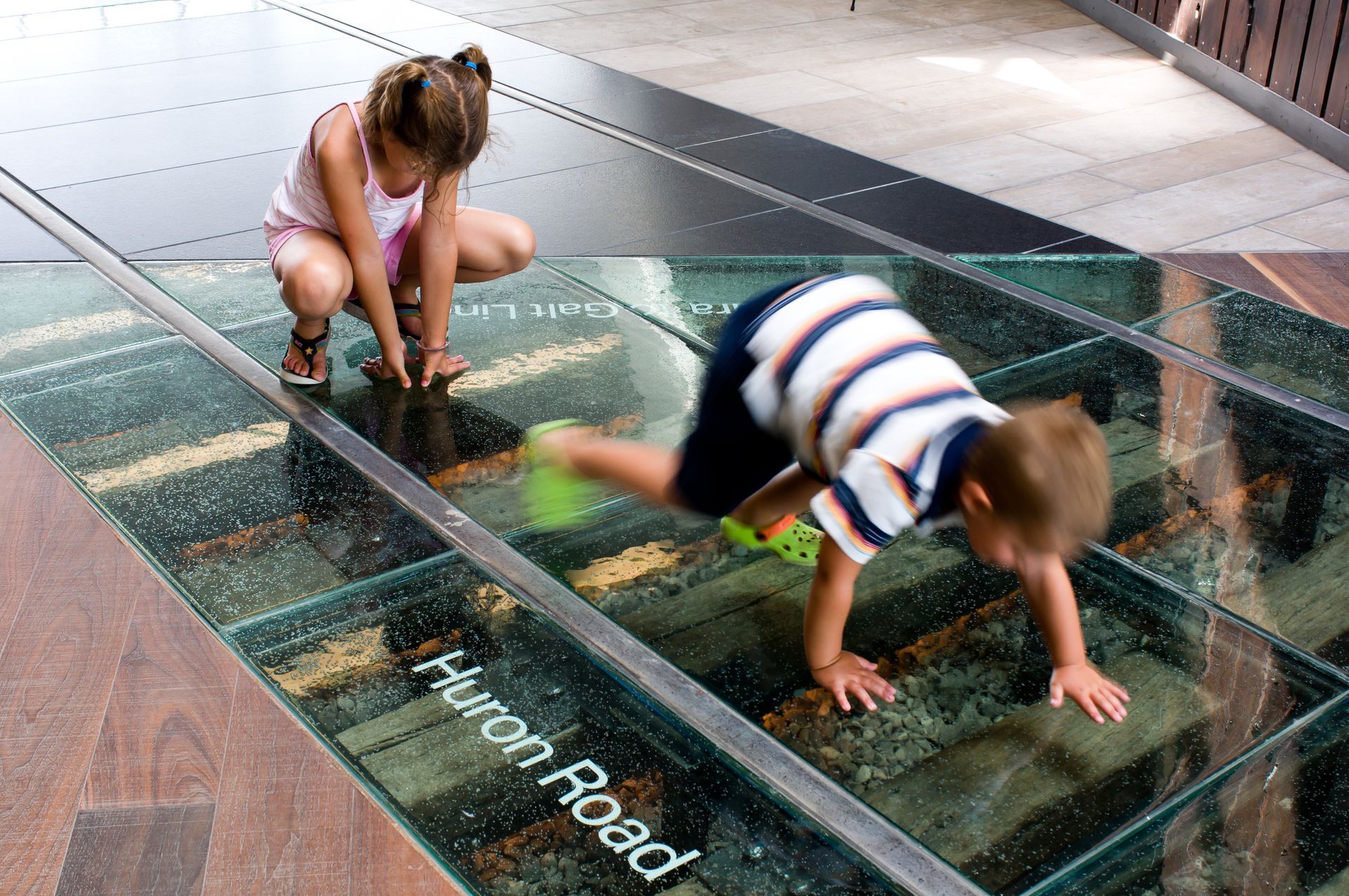 Two children playing on a glass floor section of a street revealing train tracks below.