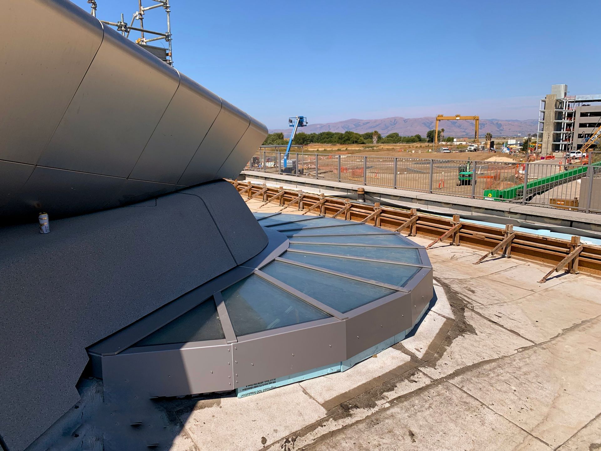 Modern building under construction with curved metal facade, glass flooring panels, and construction site in background.