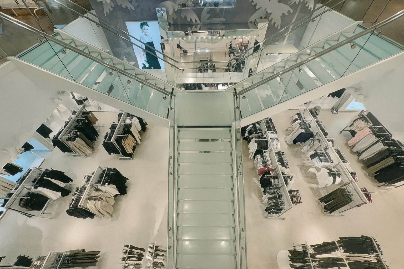 High-angle view of a multi-level clothing store with racks of clothes and shoppers. White and neutral tones dominate.