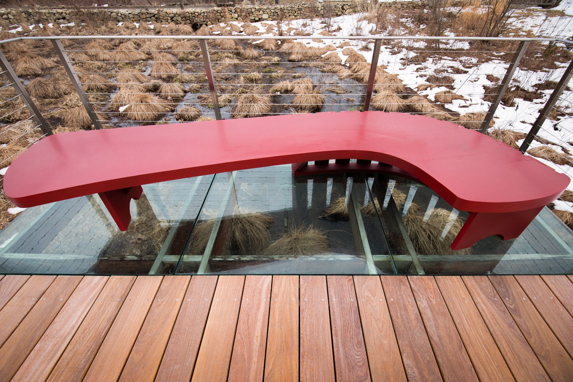 Red curved bench on a glass floor above tall, dried grass and a wooden deck. Snowy background.