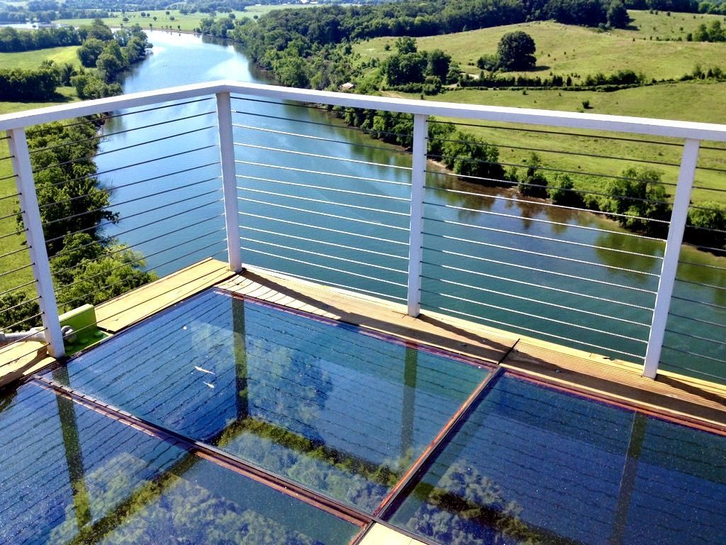 Glass-floored viewing platform overlooking a river and green landscape, white railing, sunny day.