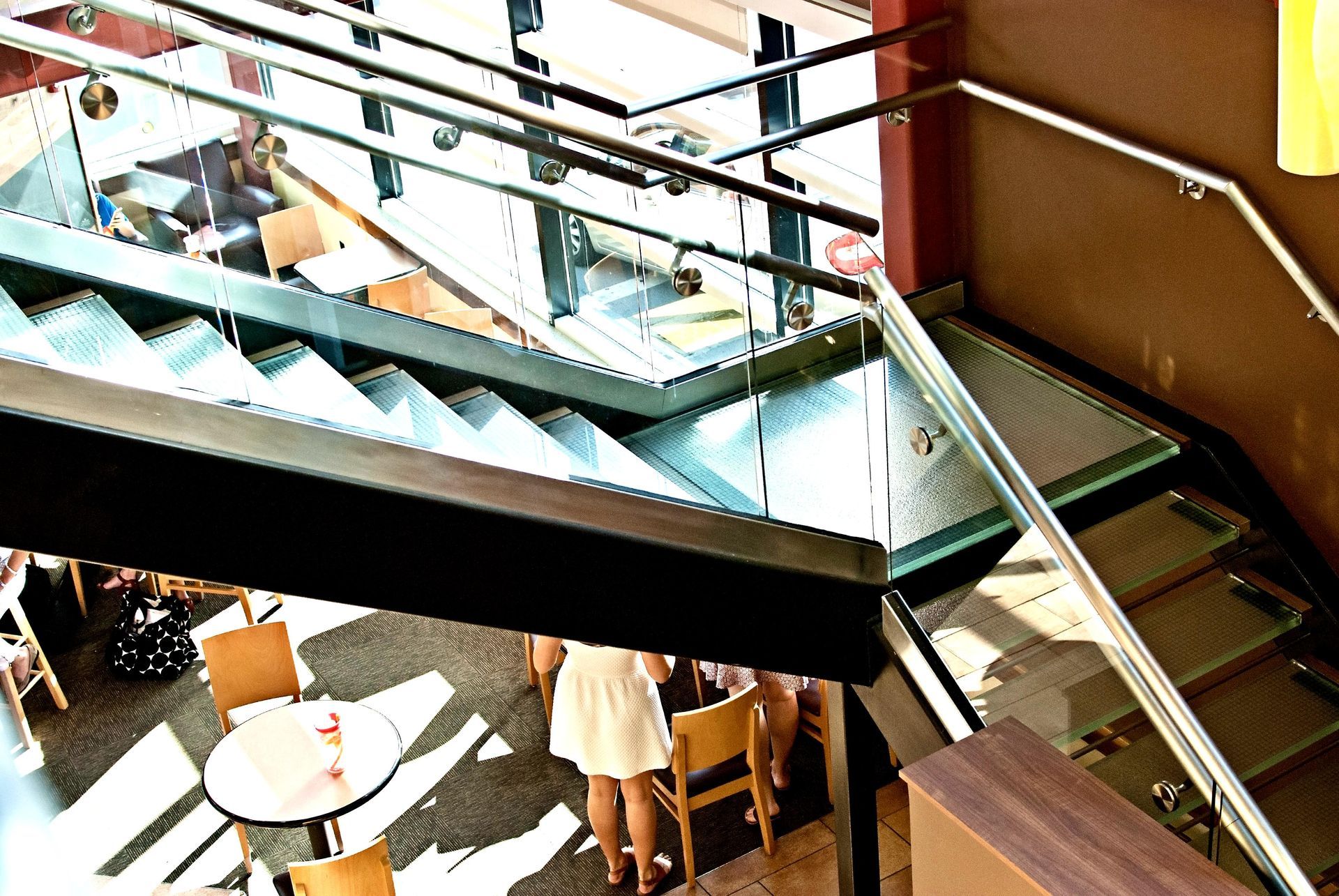 Staircase with glass railing, leading down to a cafe. People and tables are visible below.