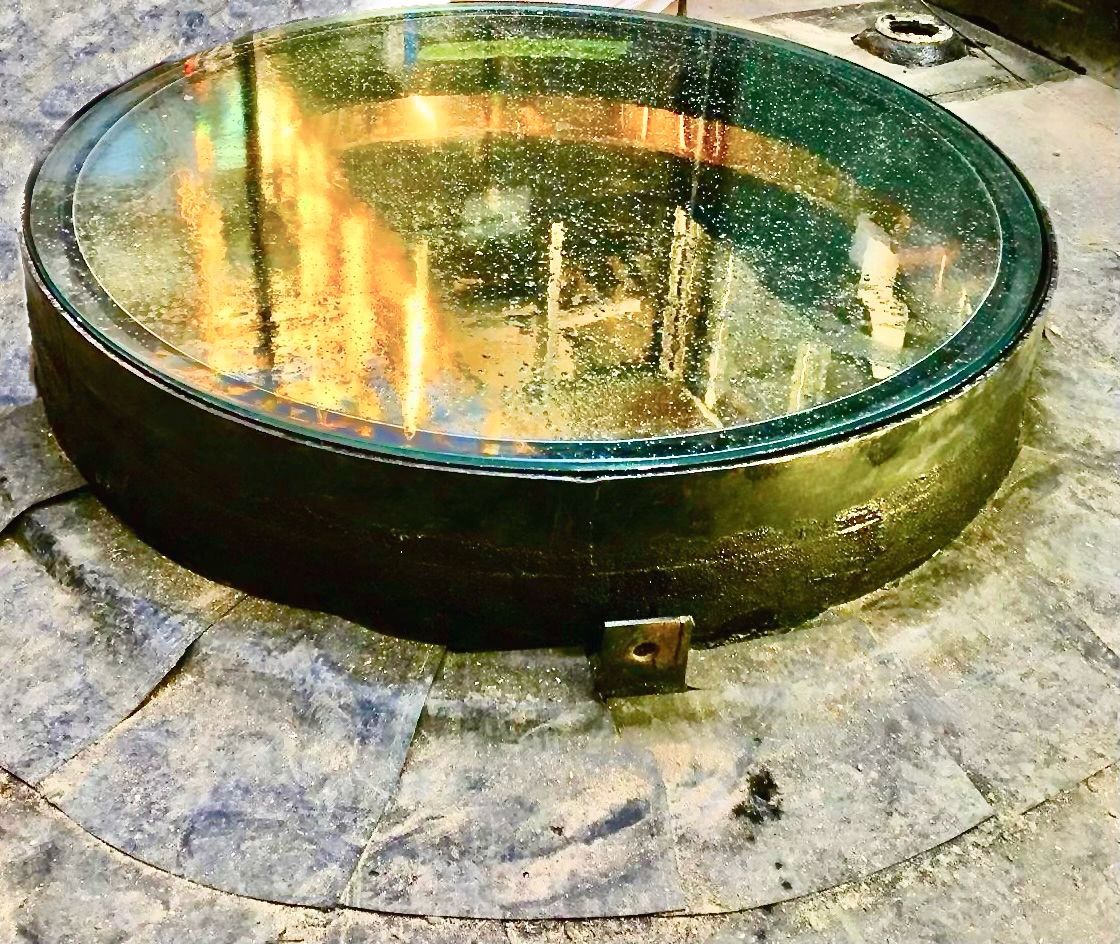 Large, round glass floor with metal rim. Appears set in a grey, dusty surface with metal bracket attached.