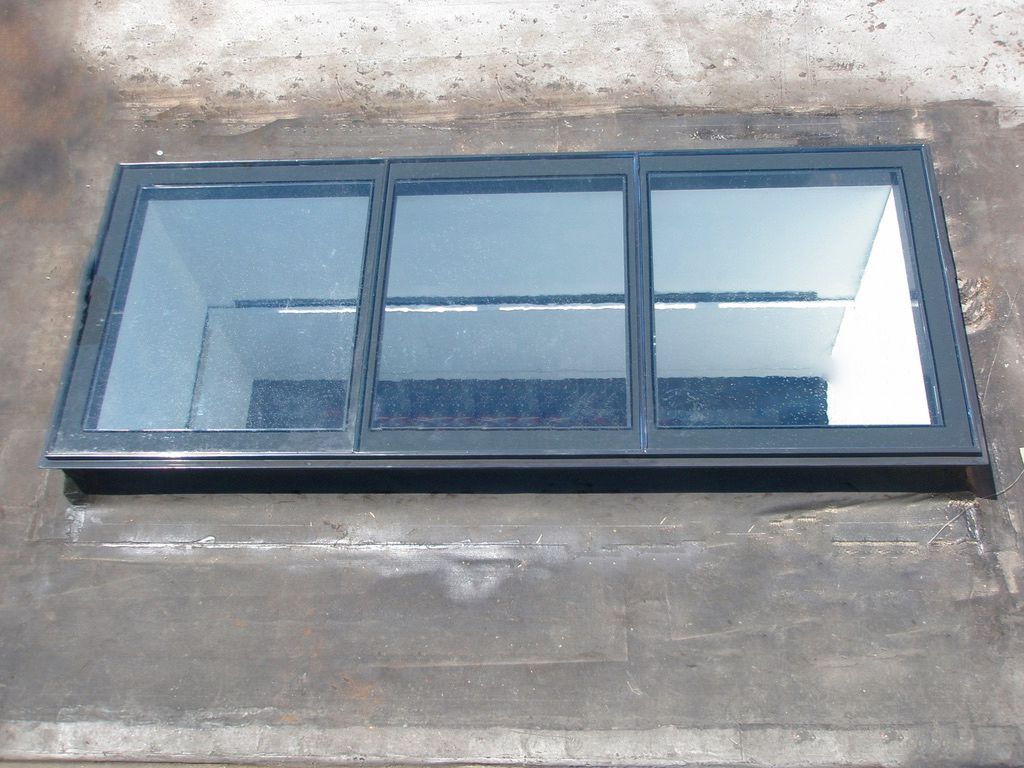 A rectangular skylight with three glass flooring panels in a dark frame on a gray roof.