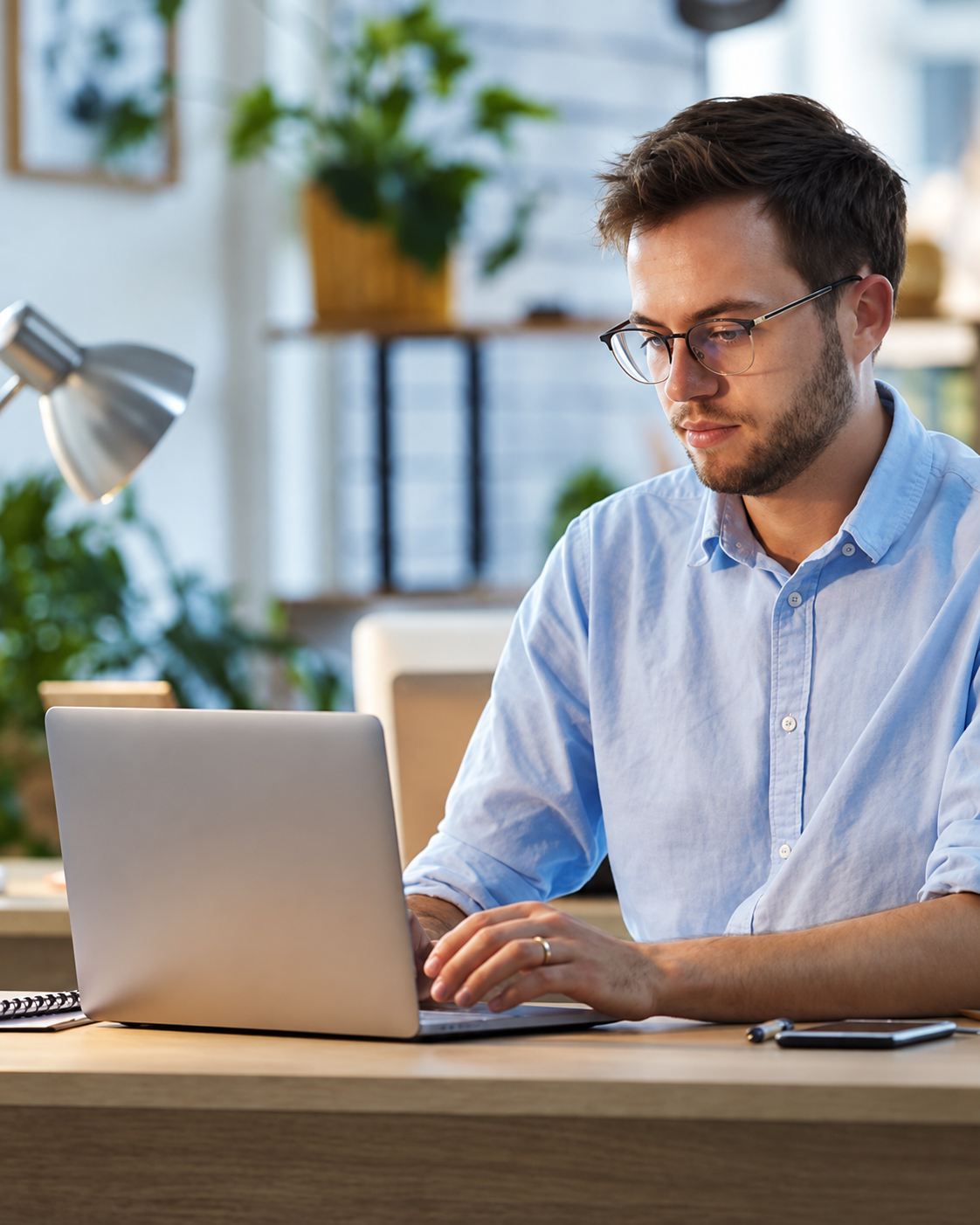 Persona trabajando en un ordenador portátil en un escritorio de una oficina luminosa, con gafas y camisa azul claro.