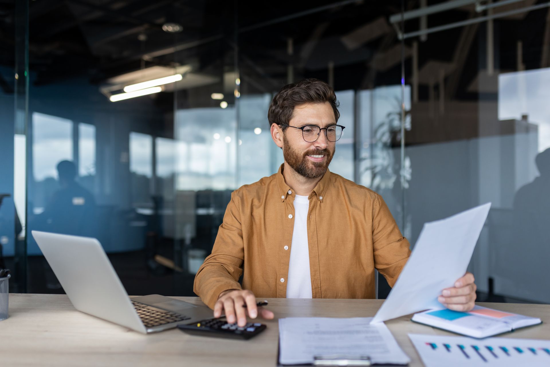 Man at office desk reviewing papers beside an open laptop in a glass-walled workspace