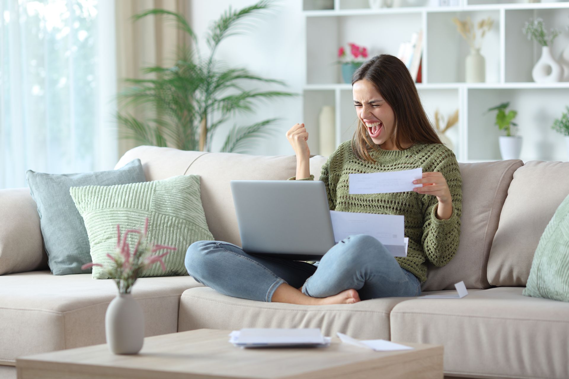 Woman sitting on a sofa, holding a letter and celebrating beside a laptop in a bright living room Woman sitting on a sofa, holding a letter and celebrating beside a laptop in a bright living room
