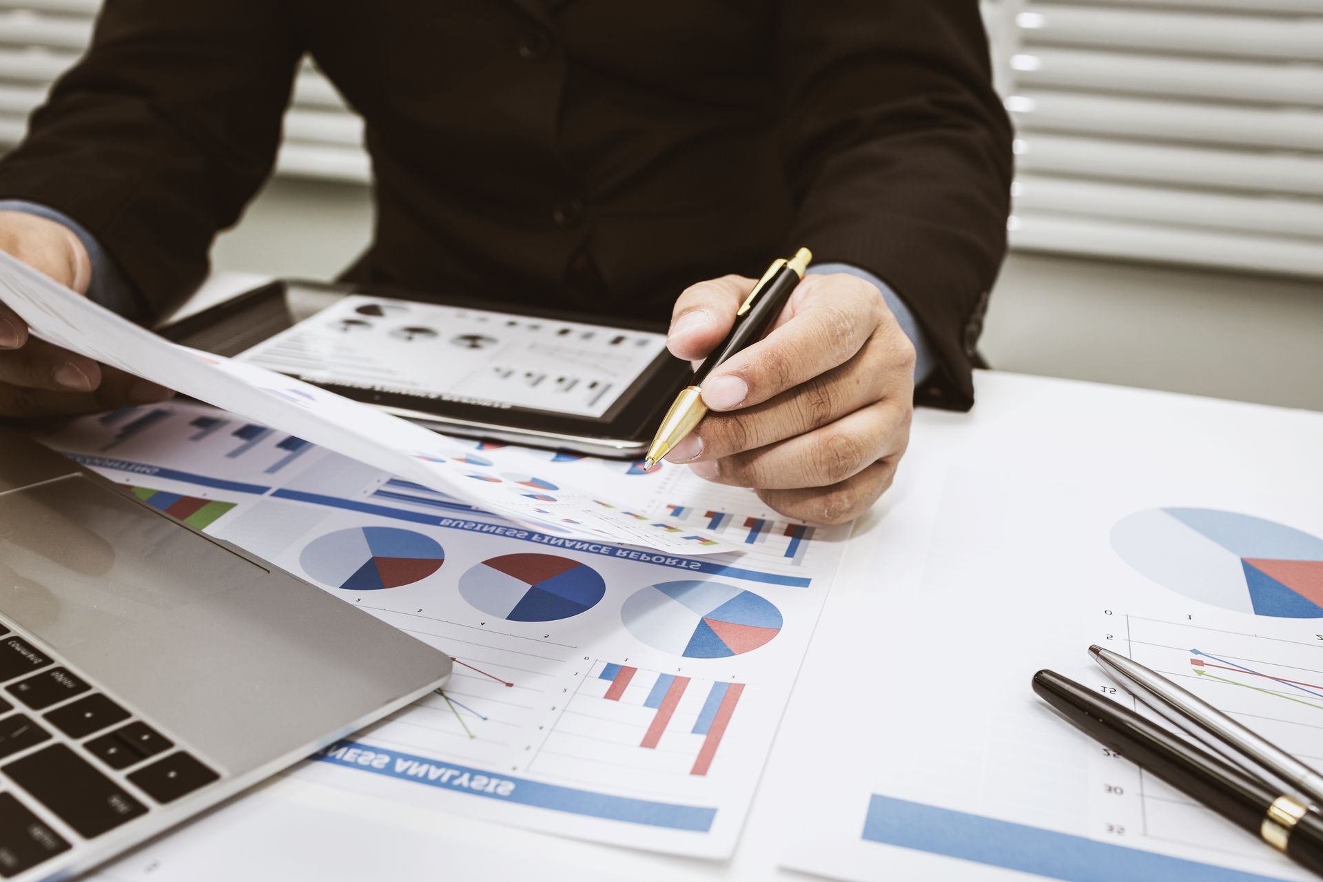 Person reviewing charts and documents beside a laptop at a desk, holding a pen. Person reviewing charts and documents beside a laptop at a desk, holding a pen.