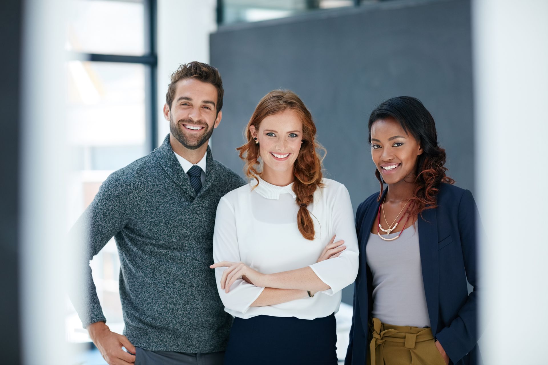 Three coworkers smiling in a bright office hallway, standing close together with arms crossed. Three coworkers smiling in a bright office hallway, standing close together with arms crossed.