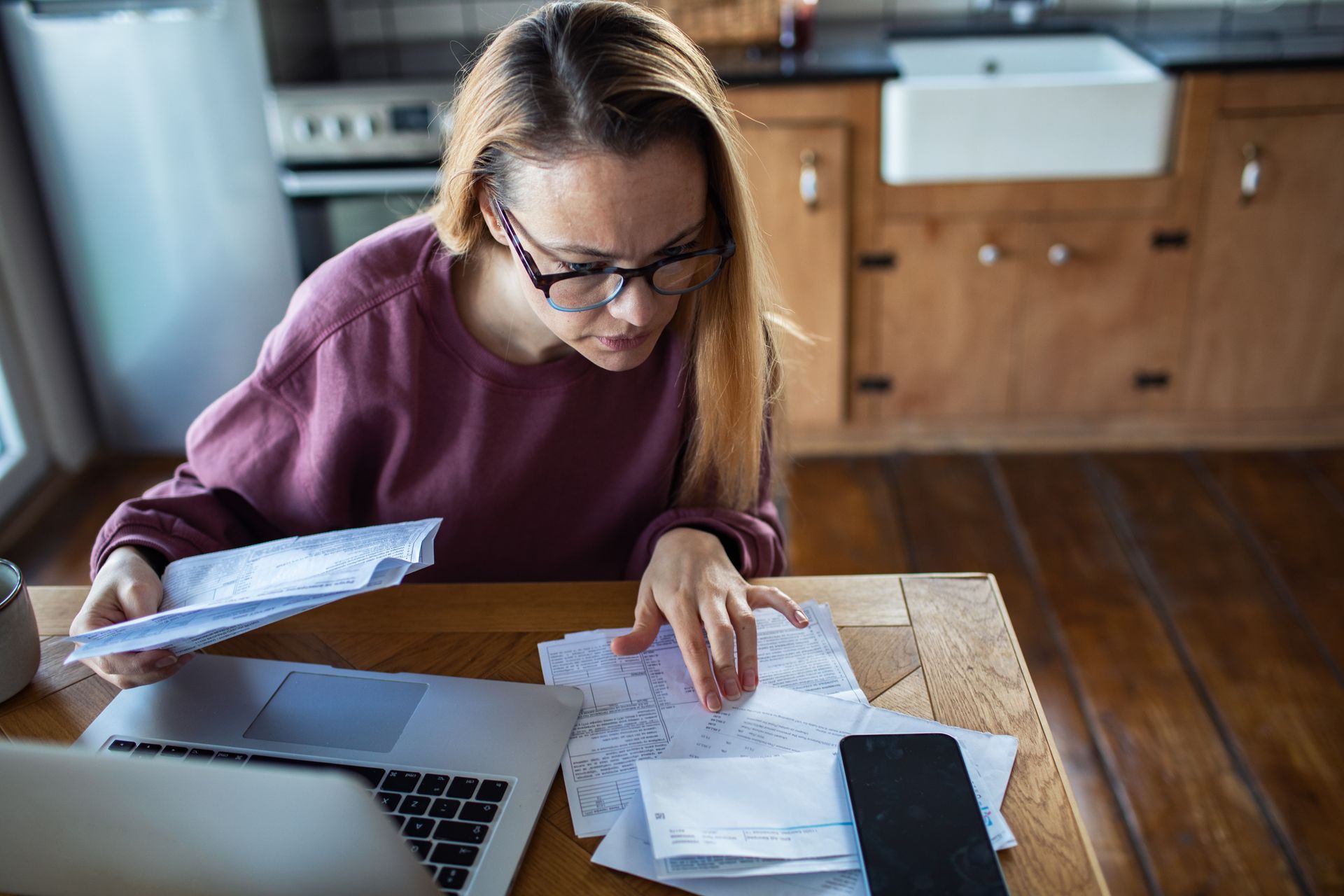 Person at a kitchen table reviewing papers and a laptop, wearing glasses and a maroon sweater. Person at a kitchen table reviewing papers and a laptop, wearing glasses and a maroon sweater.