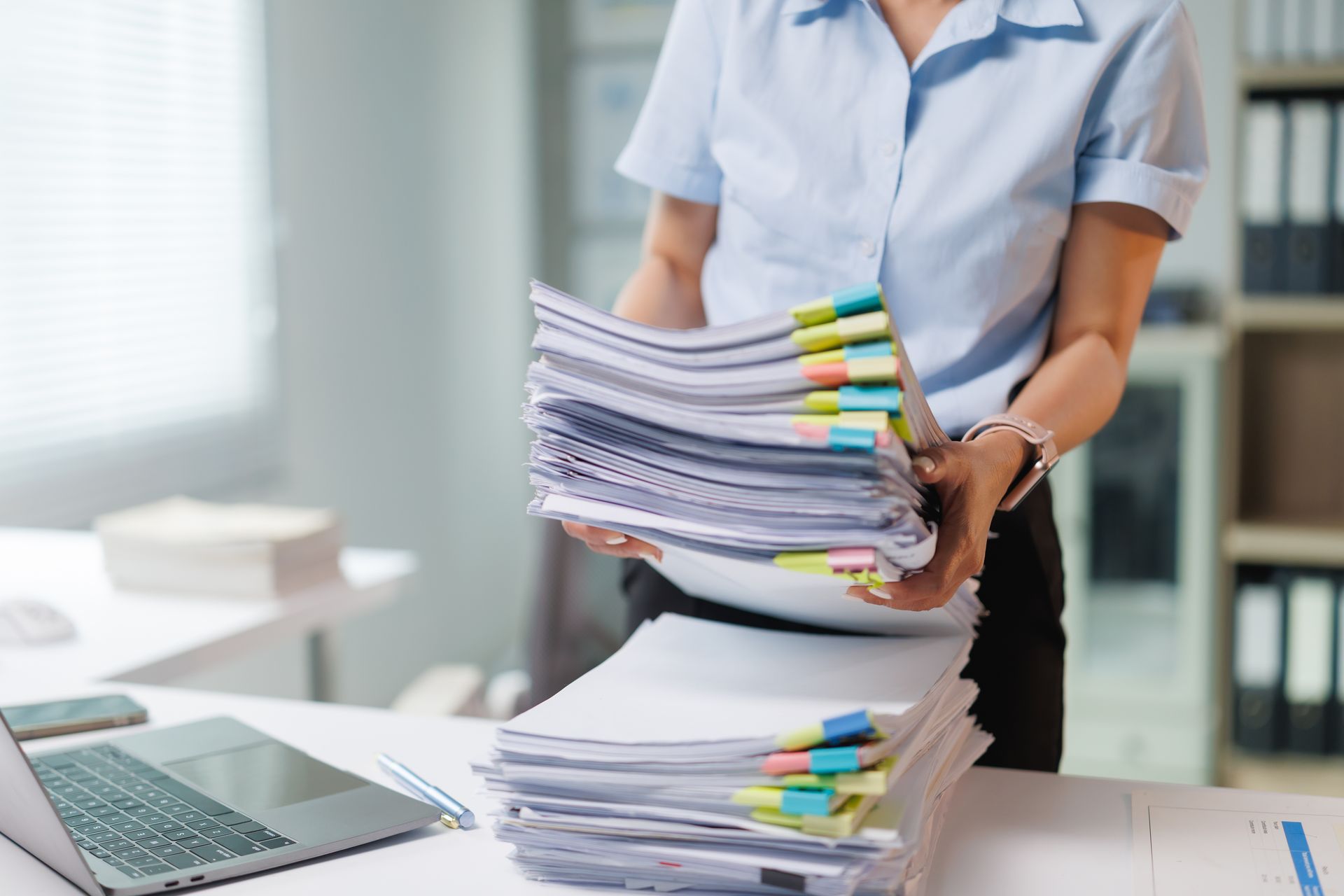 Person placing stacks of color-tabbed documents on an office desk beside a laptop Person placing stacks of color-tabbed documents on an office desk beside a laptop