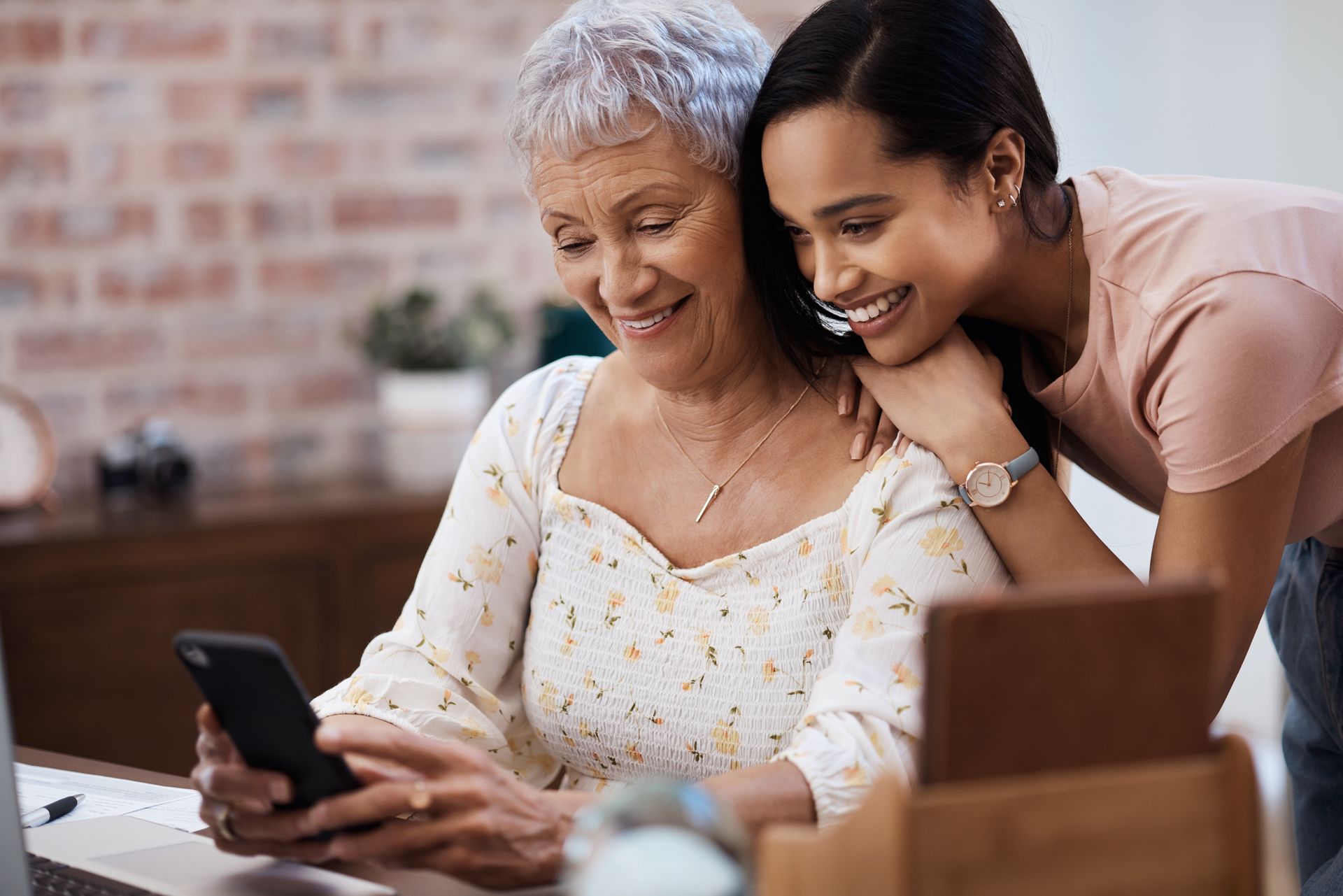 Two women smiling at a table while looking at a smartphone indoors Two women smiling at a table while looking at a smartphone indoors
