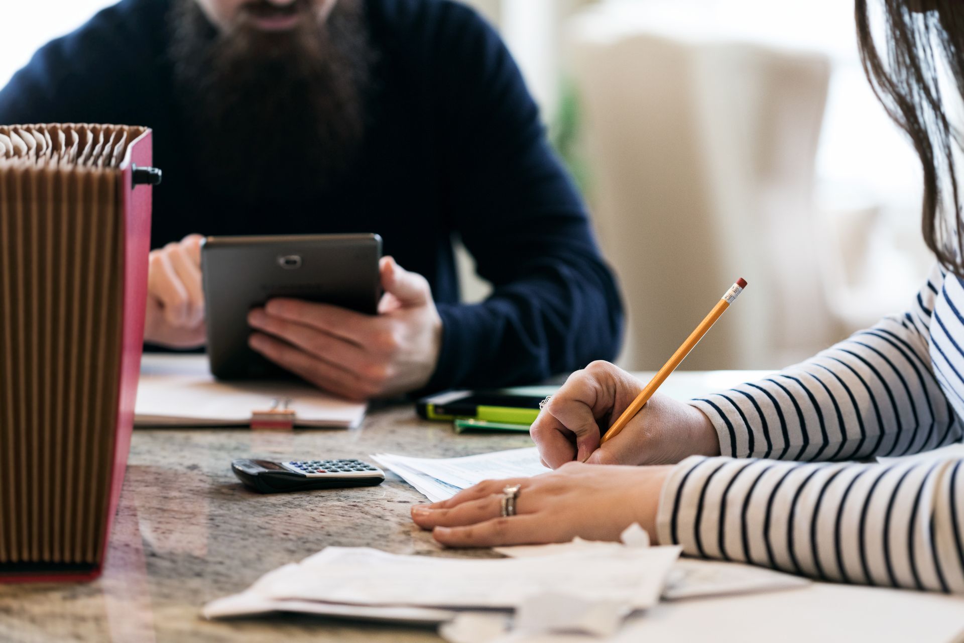 People writing and using a phone at a table with papers, pens, and a folder People writing and using a phone at a table with papers, pens, and a folder