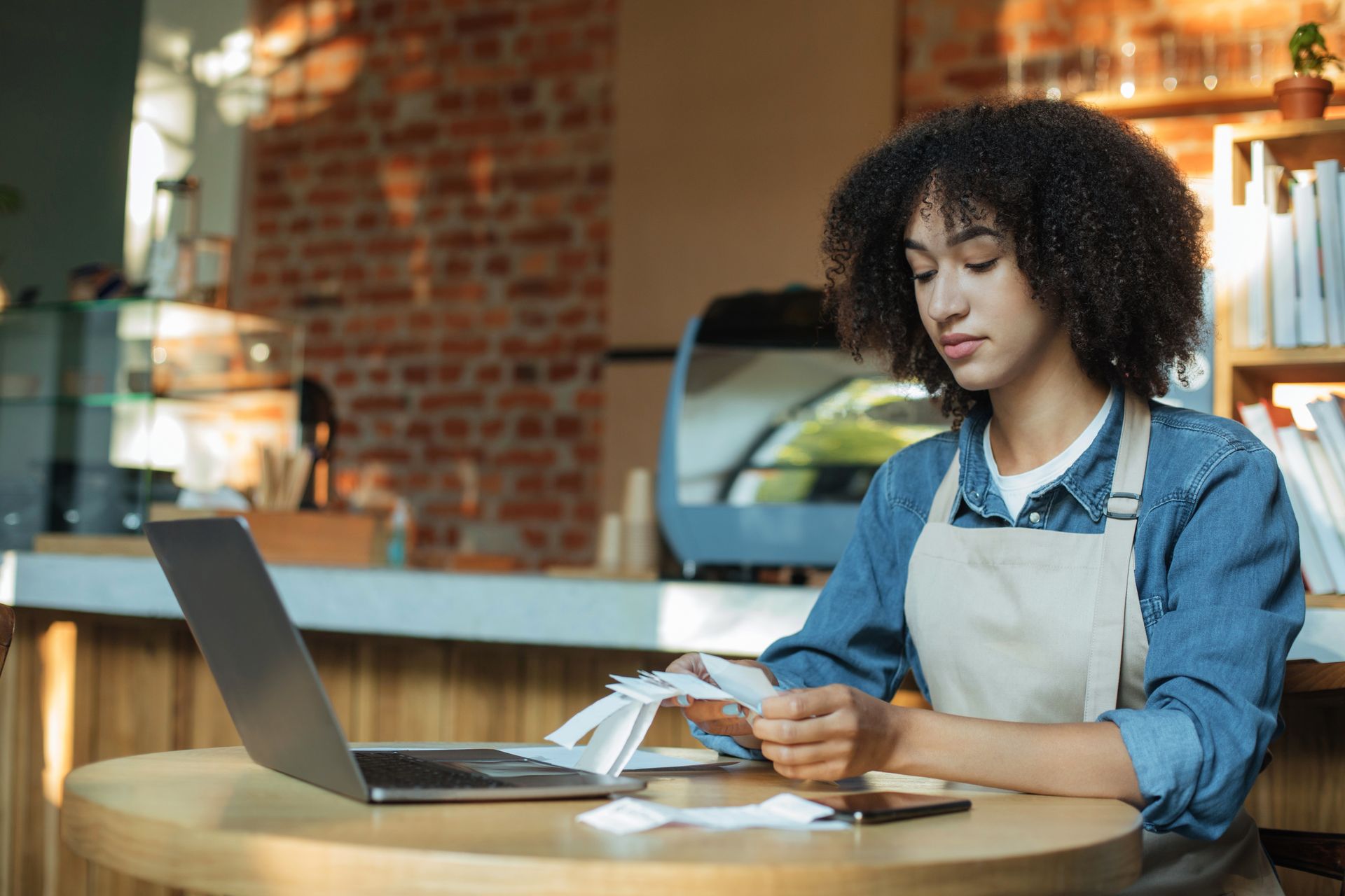 Person using a laptop and checking a receipt at a café table with brick walls and warm lights. Person using a laptop and checking a receipt at a café table with brick walls and warm lights.