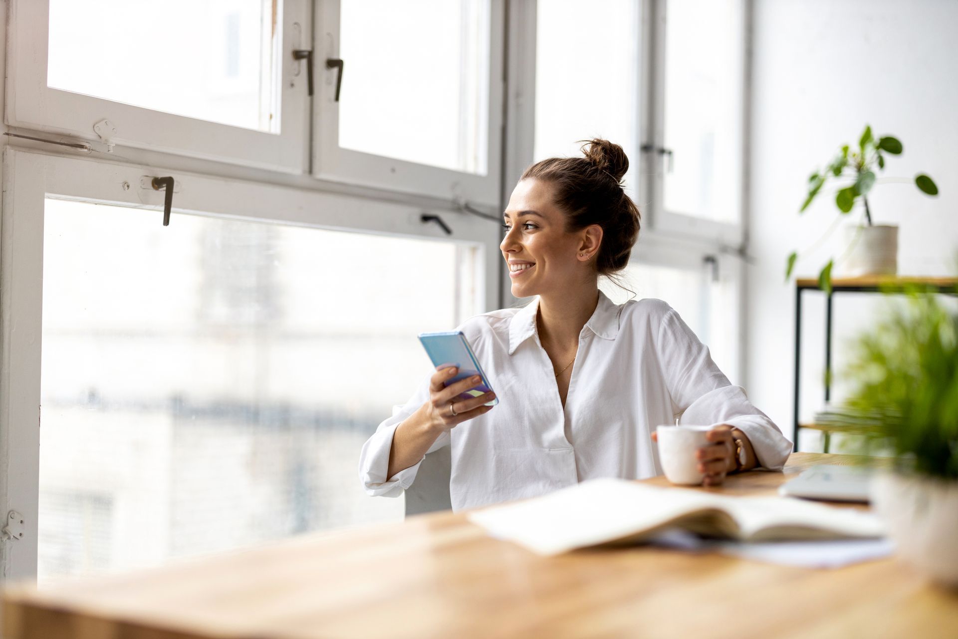 Smiling person in a bright kitchen holding a mug and phone by a window Smiling person in a bright kitchen holding a mug and phone by a window