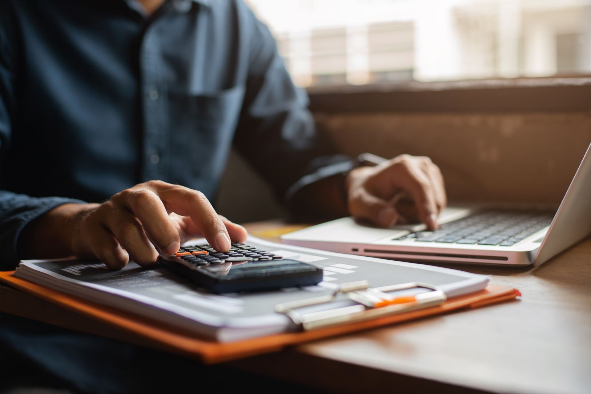 Person working at a desk with a calculator, notebook, and laptop in warm sunlight. Person working at a desk with a calculator, notebook, and laptop in warm sunlight.