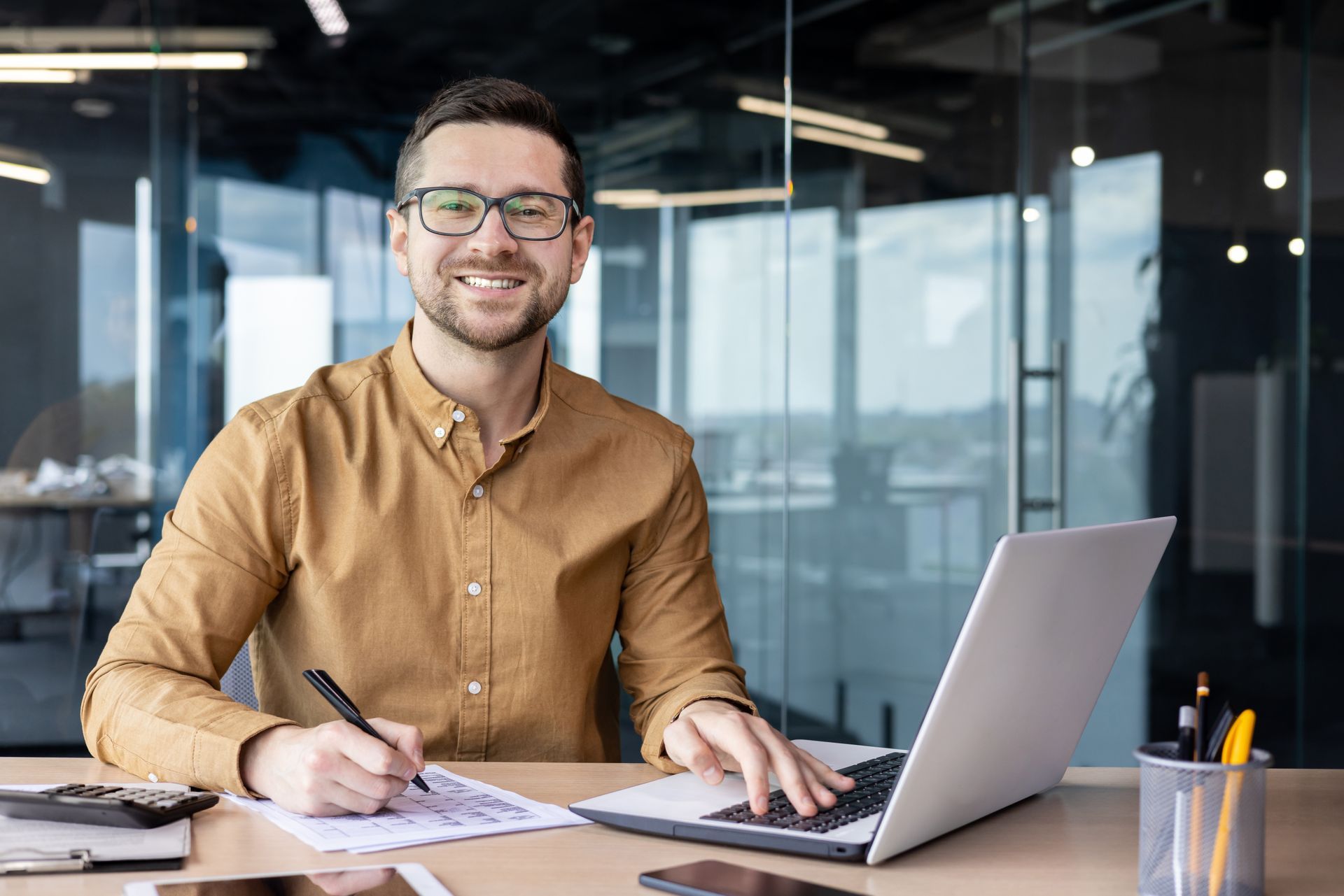 Man sitting at a desk with a laptop, papers, and pen in a glass-walled office, smiling at the camera