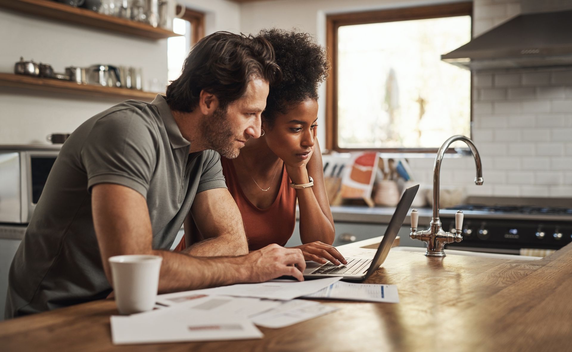 Two people leaning over a laptop at a kitchen table, reviewing papers in a bright kitchen. Two people leaning over a laptop at a kitchen table, reviewing papers in a bright kitchen.