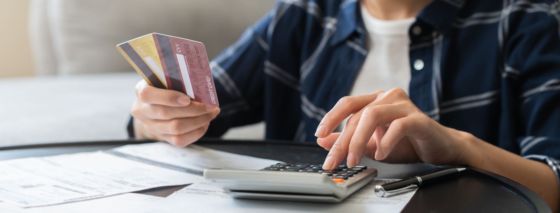 Person holding credit cards and using a calculator at a desk with paperwork and a laptop Person holding credit cards and using a calculator at a desk with paperwork and a laptop