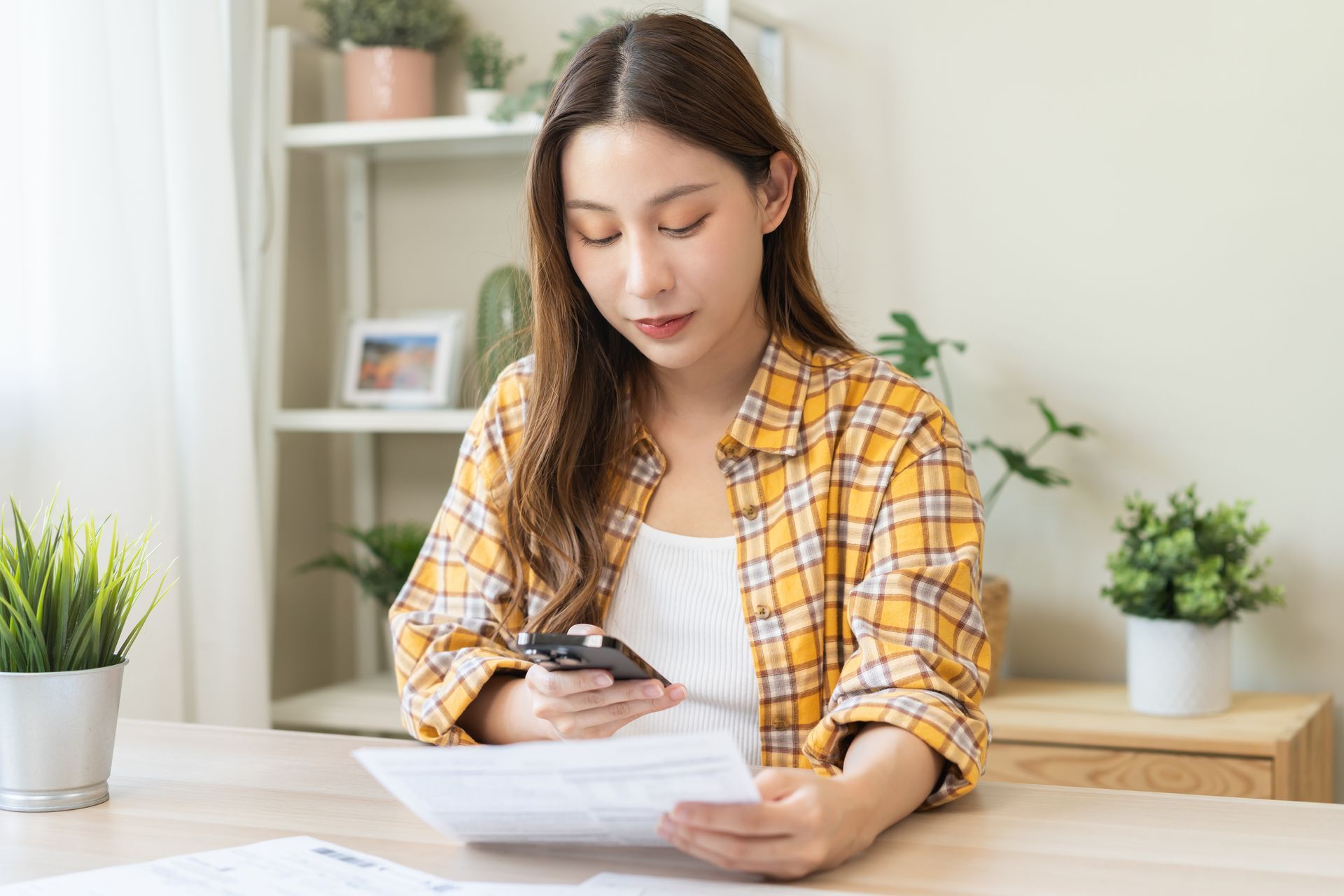 Woman in a yellow plaid shirt reads a document while checking her phone at a desk in a bright home office. Woman in a yellow plaid shirt reads a document while checking her phone at a desk in a bright home office.